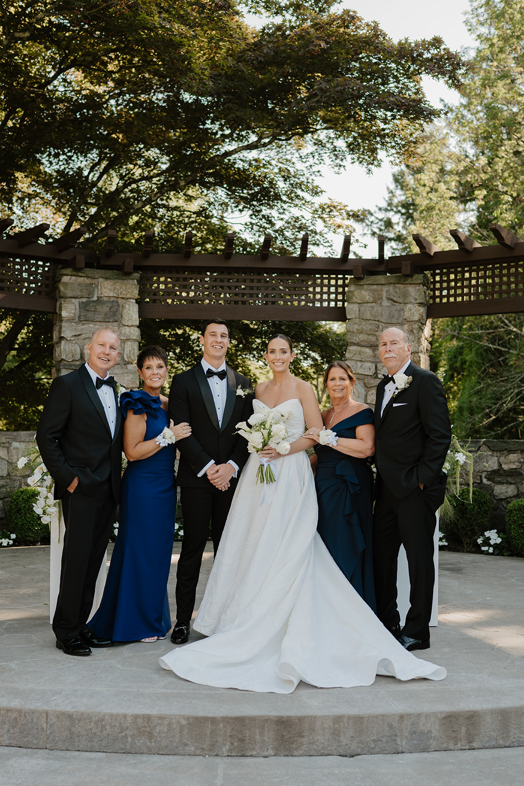 Bride and groom standing proudly with both sets of parents under a wooden arbor at Le Chateau.